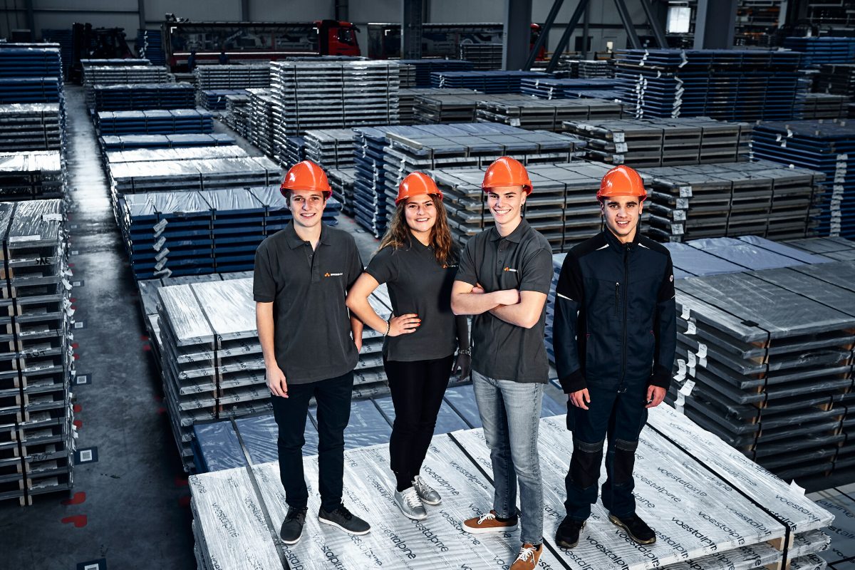 Vier Auszubildende mit orangen Schutzhelmen stehen lächelnd im Blechlager von Bucher Stahlhandel, umgeben von gestapelten Stahlblechen.
